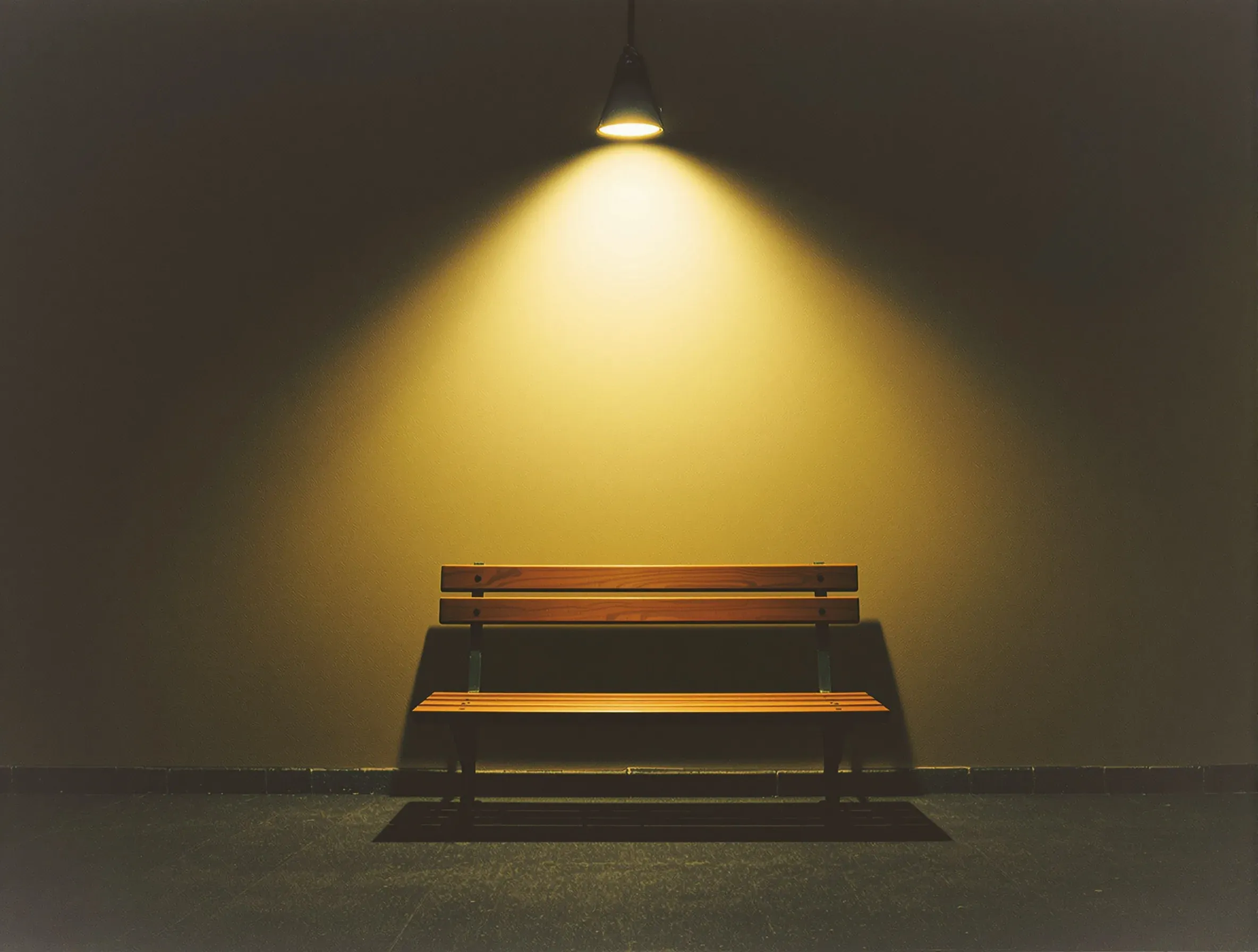 An empty bench against a bare wall under one hard overhead light