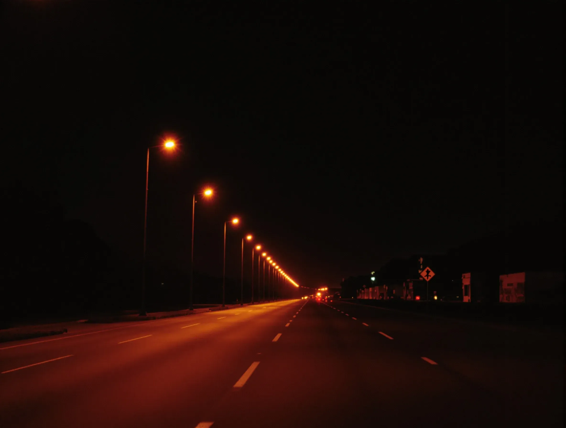 A straight two lane road at night receding under orange street lamps